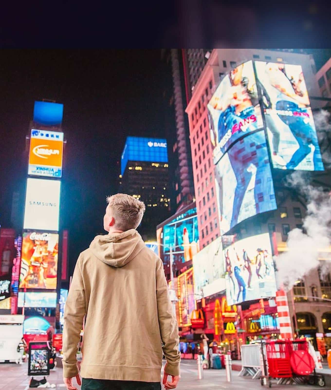 Person in a hoodie stands looking up at bright billboards and advertisements at night in Times Square, New York City.
