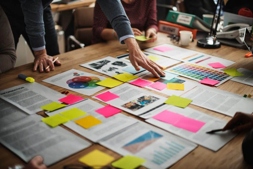 A group of people around a table filled with documents, colorful sticky notes, and diagrams, actively involved in a collaborative meeting or brainstorming session discussing why branding is important. An arm reaches over the papers.